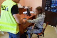 Na zdjęciu widać pomieszczenie komendy, w którym przy biurku siedzi kobieta, a obok niej stoi policjant, który zdejmuje jej kajdanki.
