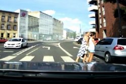 Zdjęcie przedstawia widok przez przednią szybę, z wnętrza samochodu. Widać drogę, po której jadą samochody i oznakowane przejście dla pieszych, po którym przechodzą dwie kobiety.