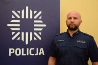 Na zdjęciu widać dzielnicowego młodszego aspiranta Grzegorza Szeligę, który stoi obok policyjnego baneru. Na banerze widać policyjną gwiazdę, pod którą widnieje napis POLICJA