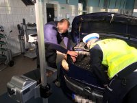 Na zdjęciu widać samochód osobowy z otwartą maską i włączonymi światłami, których ustawienie, specjalnym urządzeniem, sprawdza pracownik stacji. Czynność obserwuje policjant z Wydziału Ruchu Drogowego.