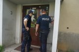 Na fotografii widać dwóch policjantów, którzy stoją przed wejściem do klatki schodowej bloku. Jeden z policjantów dzwoni na domofon.
