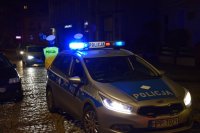 Na zdjęciu widać policyjny radiowóz z włączonymi sygnałami świetlnymi. Za radiowozem stoją dwaj policjanci z drogówki, którzy obserwują przejeżdżające samochody.