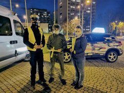 Na zdjęciu widać policjanta z drogówki, któremu towarzyszą kobieta i mężczyzna trzymający w ręku odblaski. W tle zdjęcia widać bloki mieszkalne i zaparkowane samochody.