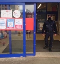 Na zdjęciu widać policjanta, który wychodzi ze sklepu wielopowierzchniowego. Na drzwiach sklepu naklejone są informacje dotyczące stosowania dystansu oraz maseczek