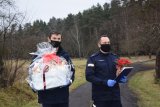 Na zdjęciu widać dwóch policjantów. Jeden z nich niesie dużą paczkę - prezent, a drugi kwiatka i list gratulacyjny. W tle zdjęcia widać rozpościerający się las.