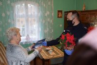 Na zdjęciu widać pomieszczenie, w którym kobieta odbiera od policjanta list gratulacyjny oraz kwiatka.