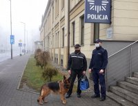 Na zdjęciu widać sierżanta sztabowego Roberta Judę oraz seniora, którzy stoją przed budynkiem Komendy Powiatowej Policji w Tarnowskich Górach. Obok nich znajduje się owczarek niemiecki Emma.