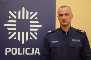 Zdjęcie przedstawia wizerunek dzielnicowego sierż. szt.  Przemysława Lebka, który stoi obok baneru. Na banerze, na niebieskim tle, widać policyjną gwiazdę, pod którą widnieje napis POLICJA