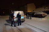Na zdjęciu widać dwóch policjantów stojących obok radiowozu. W ich kierunku zmierza bezdomny prowadzony przez policjanta i pracownika socjalnego. Mężczyzna porusza się o lasce.