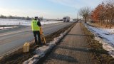 Na zdjęciu widać drogę, przy której policjant wykonuje pomiary w związku z wypadkiem. W tle zdjęcia widać samochód ciężarowy