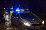 Na zdjęciu widać policyjny radiowóz z włączonymi sygnałami świetlnymi, za którym stoją, odwróceni tyłem policjanci z wydziału ruchu drogowego.