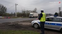 Na fotografii widać policjanta stojącego obok radiowozu. Mundurowy w dłoni trzyma tarczę służącą do zatrzymywania pojazdów. W tle fotografii widać jadący ulicą samochód ciężarowy.