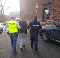 Fotografia kolorowa. Dwóch policjantów prowadzi zatrzymanego mężczyznę.