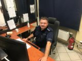 Zdjęcie kolorowe. Na fotografii widać siedzącego za biurkiem zastępcę dyżurnego aspiranta sztabowego Andrzeja Makielaka.
