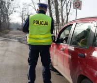 Zdjęcie kolorowe. Na fotografii widać odwróconego tyłem policjanta z ruchu drogowego, który stoi obok czerwonego samochodu osobowego.