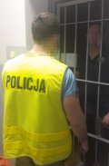Zdjęcie kolorowe. Na fotografii widać odwróconego tyłem policjanta, który obserwuje mężczyznę znajdującego się za kratami.