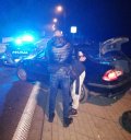 Na zdjęciu widać policjanta, który prowadzi zatrzymanego mężczyznę. Obok nich znajduje się bmw z otwartą tylną klapą oraz policyjny radiowóz z włączonymi sygnałami świetlnymi.