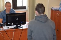 Na zdjęciu widać siedzącego za biurkiem policjanta, który przesłuchuje jednego z zatrzymanych mężczyzn.