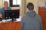 Na zdjęciu widać siedzącego za biurkiem policjanta, który przesłuchuje jednego z zatrzymanych mężczyzn.