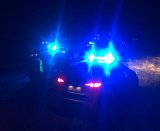 Na zdjęciu widać policyjne radiowozy z włączonymi sygnałami świetlnymi, obok których stoją policjanci.