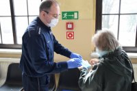 Na zdjęciu widać policjanta w maseczce i rękawiczkach jednorazowych, który przekazuje kobiecie maseczkę.