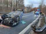 Na zdjęciu widać leżący na dachu samochód. Za samochodem widać nieoznakowany policyjny radiowóz oraz wóz strażacki.