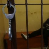 Na zdjęciu widać kajdanki zapięte na kracie w policyjnej celi, za którą siedzi zatrzymany mężczyzna.