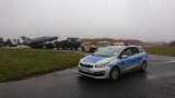 Na zdjęciu widać policyjny radiowóz z włączonymi sygnałami świetlnymi, za którym jadą inne pojazdy przewożące samolot wojskowy.