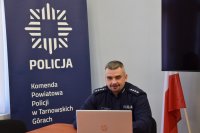 Na zdjęciu widać policyjnego profilaktyka, który siedzi przy biurku przed laptopem i prowadzi zajęcia z uczniami.