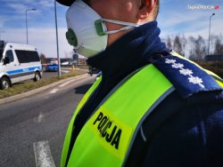 Na zdjęciu widać policjanta z białą maseczką na twarzy. W tle zdjęcia widać policyjne radiowozy.