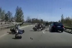 Zdjęcie przedstawia drogę, na której widać wywrócony motocykl, leżącego motocyklistę i pasażerkę. Obok nich przejeżdża samochód osobowy.