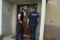 Na fotografii widać dwóch policjantów, którzy stoją przed wejściem do klatki schodowej bloku. Jeden z policjantów dzwoni na domofon.