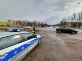 Na zdjęciu widać skrzyżowanie, przy którym znajduje się policyjny radiowóz oraz policjanci kontrolujący prędkość kierowców. Jeden z policjantów rozmawia z kierowcą samochodu zatrzymanego do kontroli drogowej.