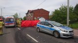 Na zdjęciu widać policyjny radiowóz, za którym znajduje się czerwony parawan. Obok parawanu widać karetkę pogotowia i strażaków.