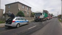 Na zdjęciu widać policyjny radiowóz, przed którym stoi zaparkowany samochód ciężarowy. Za ciężarówką widać zaparkowane wozy strażackie.