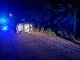 Na zdjęciu widać zniszczony samochód osobowy, który przewrócony na bok, leży na ulicy. Obok samochodu znajduje się policyjny radiowóz z włączonymi sygnałami świetlnymi.
