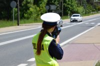 Na zdjęciu widać policjantkę z drogówki trzymającą przy twarzy urządzenie służące do pomiaru kontroli prędkości. W tle zdjęcia widać jadące ulicą samochody osobowe.