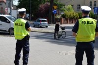 Na zdjęciu widać dwóch policjantów  z drogówki, którzy obserwują przejście dla pieszych, po którym przechodzi kobieta prowadząca rower.