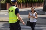 Zdjęcie przedstawia przechodzącą przez przejście dla pieszych młodą dziewczynę, która na uszach ma słuchawki, a w dłoni trzyma telefon. Po drugiej stronie przejścia czeka na nią policjant z drogówki.
