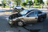 Na zdjęciu widać samochód biorący udział w wypadku. Auto jest uszkodzone i ma podniesioną maskę. W tle widać przemieszczające się osoby i samochody oraz policjantkę rozmawiającą z kobietą.