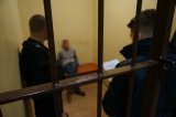 Na zdjęciu widać policyjną celę, wewnątrz której siedzi zatrzymany mężczyzna, a obok niego widać dwóch policjantów, którzy z nim rozmawiają.
