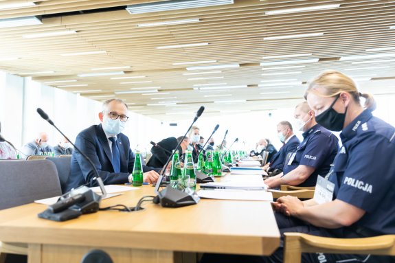 Na zdjęciu widać siedzące przy stole osoby. Wśród tych osób znajdują się policjanci. Na stole znajdują się mikrofony.