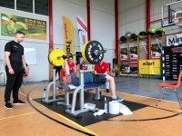 Na zdjęciu widać dzielnicowego, który leży na ławce i wyciska sztangę. Obok niego widać sędziów sportowych.