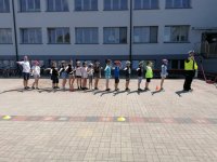 Na zdjęciu widać grupę dzieci, które mają wyciągnięte w bok prawe ręce - sygnalizując skręt w prawo. Przed dziećmi stoi policjant.