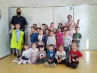 Na zdjęciu widać policjanta, który stoi obok grupy dzieci. Policjant trzyma w ręku kamizelkę odblaskową