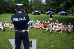 Na zdjęciu widać policjanta z drogówki, który stoi przed grupą dzieci siedzących na trawie