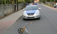 Na fotografii widać drogę, na środku której znajduje się policyjny radiowóz. Przed radiowozem, na jezdni widać przyrząd do pomiarów odległości.