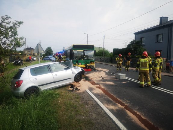 Na zdjęciu widać samochód osobowy, którego tył wpadł do przydrożnego rowu, a przód jest zgnieciony po przednią szybę. Za osobówką widać autobus, który ma uszkodzony przód i wybitą przednią szybę. W tle widać policyjny radiowóz, wozy strażackie oraz strażaków.