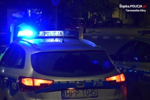 Na zdjęciu widać policyjny radiowóz z włączonymi sygnałami świetlnymi, przed którym stoją policjanci z drogówki.