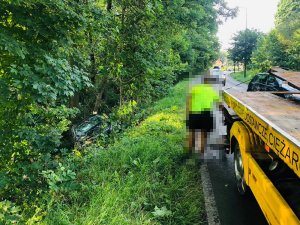 Na zdjęciu widać samochód osobowy leżący w przydrożnym rowie. Na drodze widać zaparkowaną lawetę.